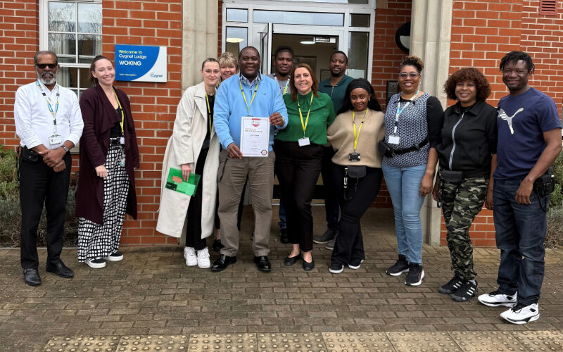 Group of diverse professionals standing in front of a brick building entrance. A person in the centre is holding a accreditation certificate.