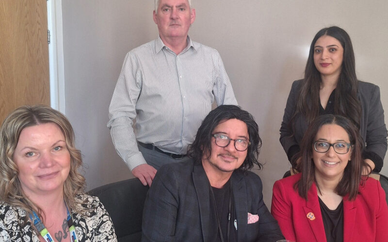 Five colleagues gathered around a wooden table. Three people seated (including a woman in a red blazer), with two others standing behind them in what appears to be an office setting