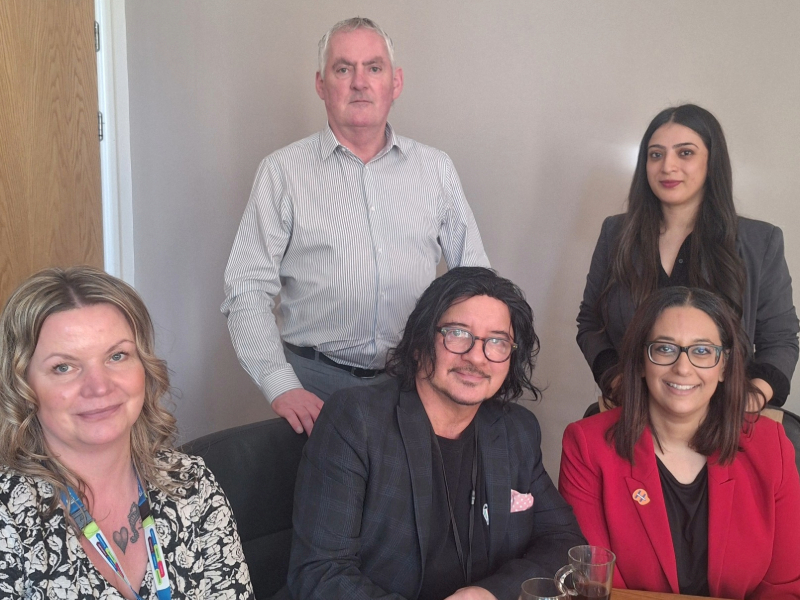 Five colleagues gathered around a wooden table. Three people seated (including a woman in a red blazer), with two others standing behind them in what appears to be an office setting