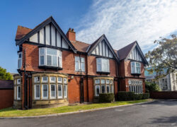 A stately Tudor-style building with distinctive architectural features. The structure has red brick walls with decorative dark timber framing on the upper levels and prominent gabled roofs. There are multiple bay windows with white frames, and the building features a large chimney