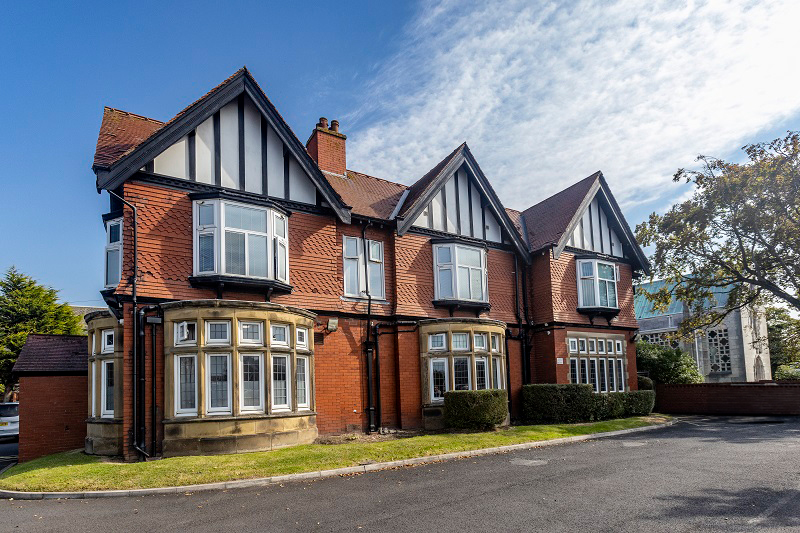 A stately Tudor-style building with distinctive architectural features. The structure has red brick walls with decorative dark timber framing on the upper levels and prominent gabled roofs. There are multiple bay windows with white frames, and the building features a large chimney