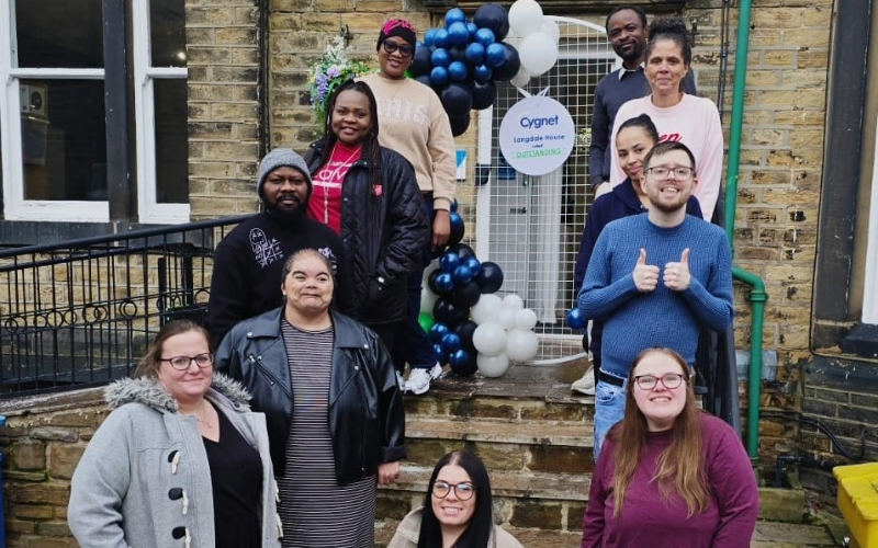A group of people standing on stone steps outside a brick building decorated with a balloon arch in dark blue and white. A circular sign on the door reads “Cygnet Langdale House Outstanding.” The setting is an entrance area with a metal gate behind the balloons and a green pipe along the wall.