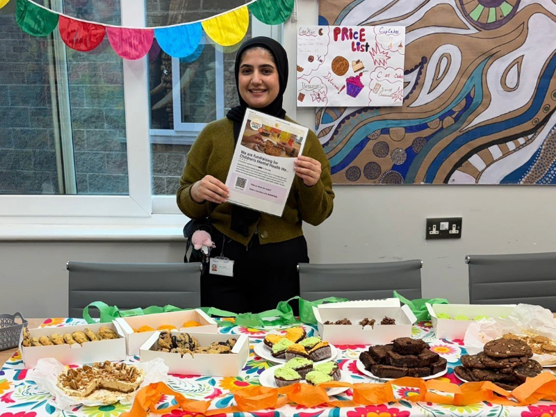 A staff member stands behind a colourful bake-sale table covered with homemade cakes, biscuits and sweet treats. The person is holding a printed fundraising poster. The table is decorated with bright bunting and colourful paper, with boxes of baked goods arranged neatly in front. Behind them, artwork and a handwritten price list are displayed on the wall.