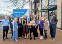 A group of staff members stand together outside Cygnet Hospital Maidstone, posing in front of a large blue sign that reads “Welcome to Cygnet Hospital Maidstone.” The team is gathered on a paved area beside the hospital building, smiling and wearing their ID lanyards. They are celebrating the hospital’s upgraded CQC rating of ‘Good’.