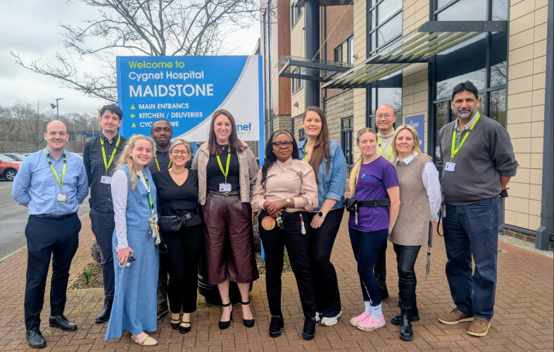 A group of staff members stand together outside Cygnet Hospital Maidstone, posing in front of a large blue sign that reads “Welcome to Cygnet Hospital Maidstone.” The team is gathered on a paved area beside the hospital building, smiling and wearing their ID lanyards. They are celebrating the hospital’s upgraded CQC rating of ‘Good’.